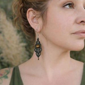 May include: A close-up of a macrame earring with a tiger's eye stone. The earring is dark green and brown, with a detailed woven pattern. The stone is a warm, golden brown color. The earring hangs from a silver hook.