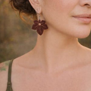 May include: A close-up of a brown, flower-shaped earring. The earring is made of woven material and hangs from a silver hook. The earring is a deep brown color and has a detailed, textured design. The earring is worn on a person's ear.