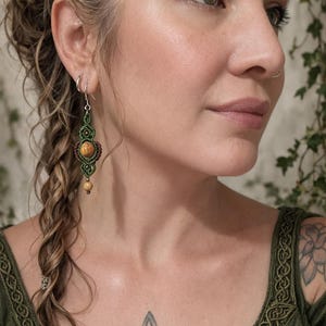May include: Handmade macrame earrings with a central amber-colored stone and green and brown accents. The earrings are suspended from silver-colored hooks. The design features intricate knotting and a bohemian aesthetic.