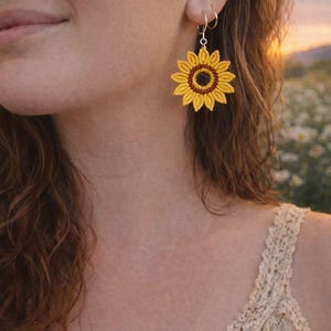 May include: Crocheted sunflower earrings with yellow petals and a brown center. The earrings are attached to silver hoops. The image shows a close-up of the earrings on a person's ear, with a blurred background of a field.