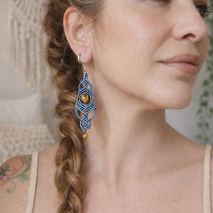 May include: A pair of blue macrame earrings with a central amber-colored stone and smaller amber beads. The earrings have a long, teardrop shape and are suspended from silver-colored ear wires. The design features intricate knotting patterns.