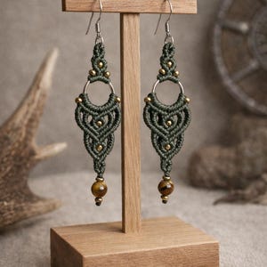 May include: A pair of handmade macrame earrings in dark green, featuring a geometric design with gold-colored beads and a round, silver-colored hoop. The earrings are displayed on a wooden stand, with a brown tiger eye bead at the bottom.