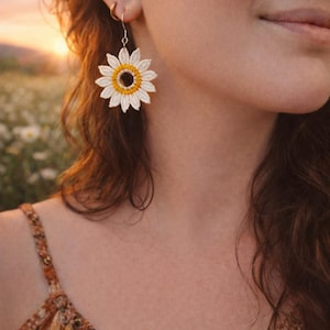 May include: A close-up of a white and yellow crochet sunflower earring. The earring is a detailed replica of a sunflower, with white petals and a yellow center. The earring is hanging from a gold hoop. The background is blurred, with a field of flowers and a sunset.