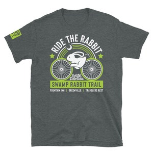 May include: A dark gray t-shirt with a green and white graphic design. The graphic features a bicycle with a rabbit riding it, a crescent moon, and the text "Ride the Rabbit, South Carolina, Swamp Rabbit Trail, Fountain Inn, Greenville, Travelers Rest".