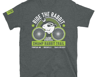 May include: A dark grey t-shirt with a green and white graphic design. The graphic features a bicycle with a rabbit riding it, a crescent moon, and the text "Ride the Rabbit, South Carolina, Swamp Rabbit Trail, Fountain Inn, Greenville, Travelers Rest".
