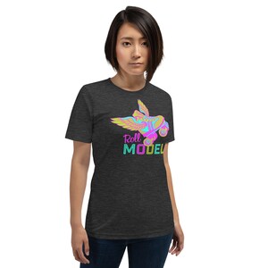 May include: A dark grey t-shirt with a vibrant graphic of a winged roller skate and the words "Roll Model". The roller skate design incorporates pink, blue, yellow, and green hues. The t-shirt has a crew neck and short sleeves.
