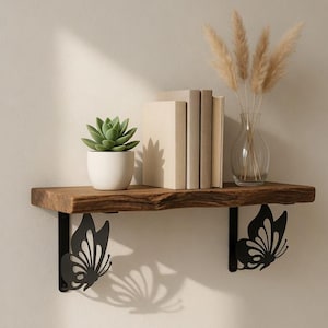 May include: A rustic wooden shelf with black metal butterfly supports. The shelf holds a small potted succulent, books, and a vase of dried flowers. The shelf is mounted on a neutral-colored wall.