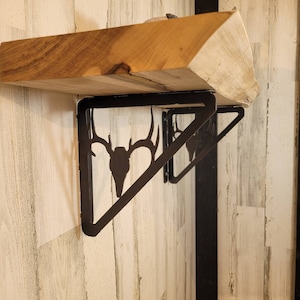 May include: A rustic wooden shelf with a natural edge, supported by black metal brackets featuring a deer skull silhouette. The shelf is made of light brown wood and is mounted on a white and gray wooden wall.