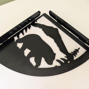 May include: A black metal corner shelf featuring a cutout silhouette of a bear walking in front of a mountain range and pine trees. The shelf has two perpendicular mounting brackets with pre-drilled holes for installation in a corner.