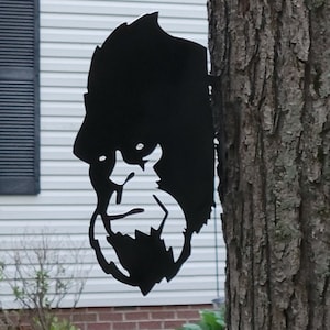 May include: A black metal silhouette of a Bigfoot's head is attached to a tree trunk. The silhouette is detailed with a beard and furrowed brow.