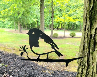May include: Black metal silhouette of a bird perched on a branch, mounted on a tree trunk. The bird is facing left and has a detailed design. The background is a blurred green lawn and trees.