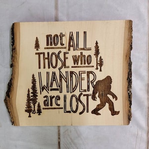 May include: Wooden sign with a rustic bark edge, featuring the quote "Not all those who wander are lost" with a silhouette of Bigfoot walking. The sign is a light brown color with dark brown lettering.