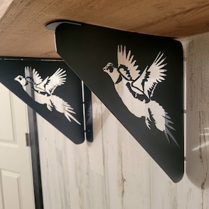 May include: Two black metal shelf brackets with a pheasant design cut out. The brackets are mounted to a wooden shelf.