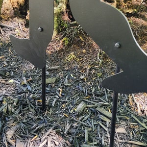 May include: Black metal silhouette of a person running, with two metal stakes inserted into the ground to hold it up.