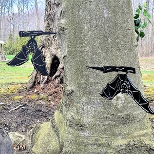 May include: Two black metal bat silhouettes are attached to a tree trunk. The bats are in a flying position with their wings spread out. The tree trunk is brown and has a rough texture.