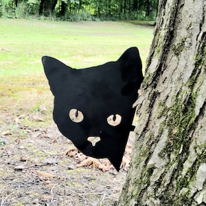 May include: Black metal silhouette of a cat peeking from behind a tree trunk. The cat has large, round eyes and a playful expression.
