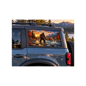May include: A gray SUV features a rear window decal depicting a brown Bigfoot standing in a river while fishing for a jumping trout. The scene includes a vibrant sunset, autumn trees, and distant mountains, all displayed on the vehicle's glass.