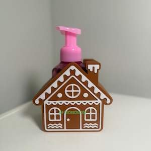 May include: A brown gingerbread house-shaped soap dispenser with white icing details. The dispenser has a pink pump and a clear bottle. The design includes windows, a door, and a chimney. The text "A TOUCHED CREATION" is visible on the house.