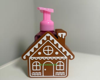 May include: A brown gingerbread house-shaped soap dispenser with white icing details. The dispenser has a pink pump and a clear bottle. The design includes windows, a door, and a chimney. The text "A TOUCHED CREATION" is visible on the house.