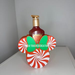 May include: A red and white peppermint-themed soap dispenser holder shaped like Mickey Mouse ears, with a green bow. The holder surrounds a brown bottle with a gold pump. The text "@A TOUCHEDCREATION" is visible.