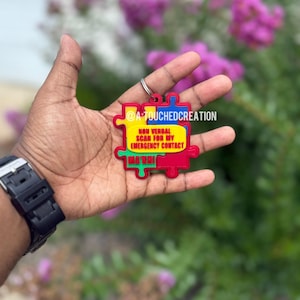 Op de afbeelding: Een kleurrijke puzzelstukje sleutelhanger met de tekst "NON VERBAL SCAN FOR MY EMERGENCY CONTACT". De sleutelhanger is rood, geel, blauw en groen. De tekst is wit. De sleutelhanger heeft een zilveren ring.