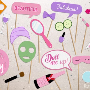 May include: A set of 14 pink and purple photo booth props with spa-themed designs. The props include a wine glass, a face mask, a headband, a pair of cucumbers, a bottle of champagne, a makeup brush, a pair of eye masks, a nail polish bottle, a lipstick, a martini glass, a mirror, a bow tie, a speech bubble that says "Fabulous!" and a speech bubble that says "Beautiful!"