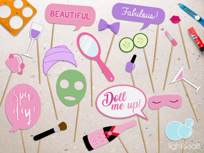 May include: A set of 14 pink and purple photo booth props with spa-themed designs. The props include a wine glass, a face mask, a headband, a pair of cucumbers, a bottle of champagne, a makeup brush, a pair of eye masks, a nail polish bottle, a lipstick, a martini glass, a mirror, a bow tie, a speech bubble that says "Fabulous!" and a speech bubble that says "Beautiful!"