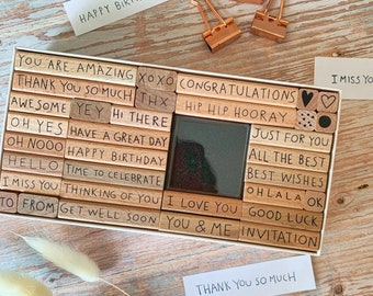 May include: A set of wooden stamps with various phrases and designs, including "Happy Birthday", "Congratulations", "I Love You", and "Just for You". The stamps are arranged in a box with a white background.