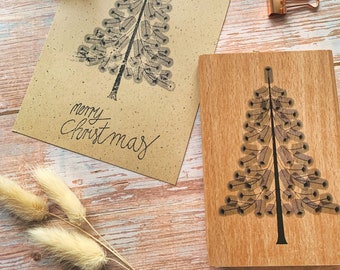 May include: A wooden stamp with a Christmas tree design and the words "merry Christmas" is shown next to a stamped card. The card is printed on brown paper. Also shown are copper binder clips and a wooden stamp handle.