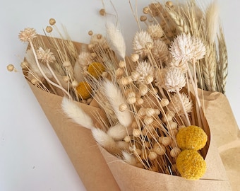 May include: A bouquet of dried flowers in shades of beige and yellow, wrapped in brown paper. The bouquet includes dried wheat, bunny tails, and small round yellow flowers.