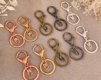 May include: Eight keychains in different metal finishes, including silver, gold, rose gold, and black. The keychains have a round ring and a lobster claw clasp.