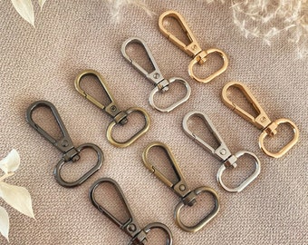 May include: Eight metal swivel clasps in different finishes: gold, silver, and black nickel. The clasps are arranged in a semi-circle on a light brown fabric background.