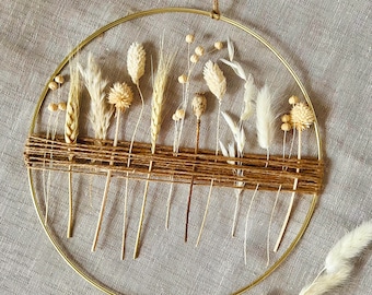 May include: A gold metal hoop with dried flowers and grasses arranged in a woven pattern. The hoop is hanging from a string and the flowers are in shades of white and beige.