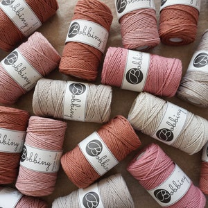 Può includere: Una collezione di bobine di spago di cotone naturale in varie tonalità di rosa, marrone e grigio. Ogni bobina è etichettata con la parola "Bobbiny".