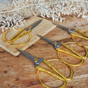 May include: Four gold-colored scissors with black handles on a wooden surface. The scissors have a decorative design on the blades.