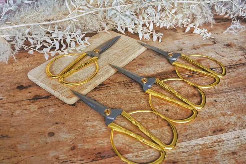 May include: Four gold-colored scissors with black handles on a wooden surface. The scissors have a decorative design on the blades.