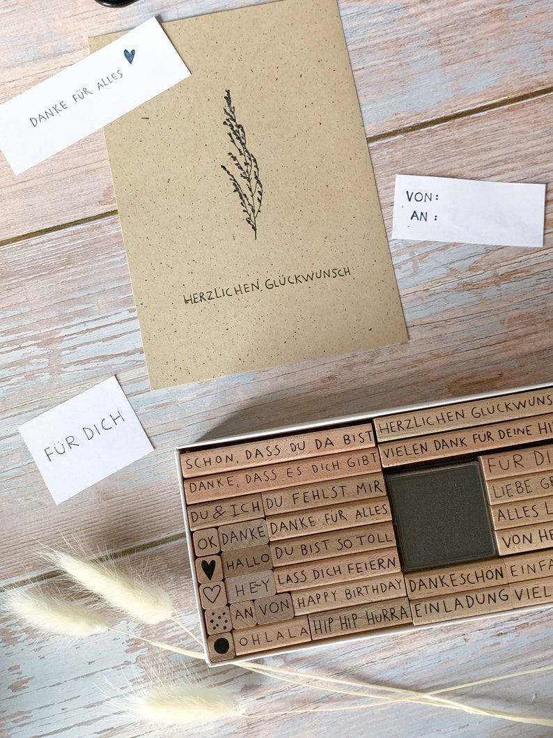 May include: A set of wooden stamps with various phrases and designs, including "Danke fur alles", "Herlichen Glueckwunsch", "Du & Ich", "Ok Danke", "Hallo", "Hey", "Lass dich feiern", "Happy Birthday", "Oh la la", "Hip Hip Hurra", "Vielen Dank", "Fur di", "Liebe ge", "Alles L", "Von He", "Dankesch&ouml;n", "Einladung", "Viel", and hearts. The stamps are arranged in a wooden box.