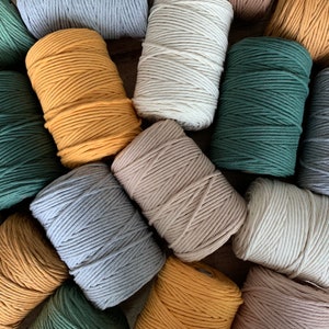 Pode incluir: Uma variedade de corda de algodão em diferentes cores, incluindo natural, bege, cinza, verde e amarelo. A corda está enrolada em bobinas e disposta sobre uma superfície de madeira.