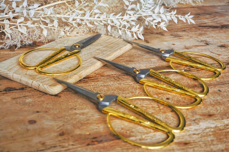 May include: Four gold-colored scissors with silver handles on a wooden surface. The scissors are arranged in a row, with the handles pointing towards the top of the image.