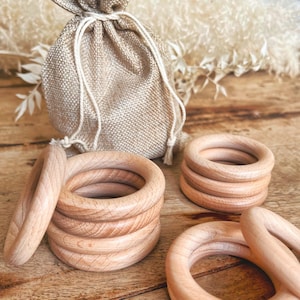 Può includere: Una collezione di anelli per la dentizione in legno in varie pile e disposizioni. Gli anelli sono realizzati in legno di colore chiaro e hanno un design liscio e arrotondato. Sullo sfondo un piccolo sacchetto di iuta con coulisse.