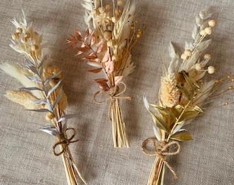 May include: Three small bouquets of dried flowers tied with twine. The bouquets are made up of dried grasses, pampas grass, and small white flowers. The bouquets are arranged on a beige linen background.