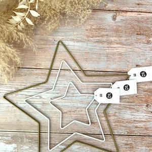 May include: Three gold wire star shapes, one inside the other, on a wood background. The largest star is green, the middle star is white, and the smallest star is white. There are three white tags with black polka dots and black text that says "Made by Me" on them.