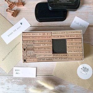 May include: A set of wooden stamps with various phrases in German, including "Danke fur alles", "Herlichen Glueckwunsch", "Du fehlst mir", and "Alles Liebe". The stamps are arranged in a rectangular box with a black ink pad. The box is on a wooden surface with two envelopes and a small white circle with a drawing of an envelope with flowers.