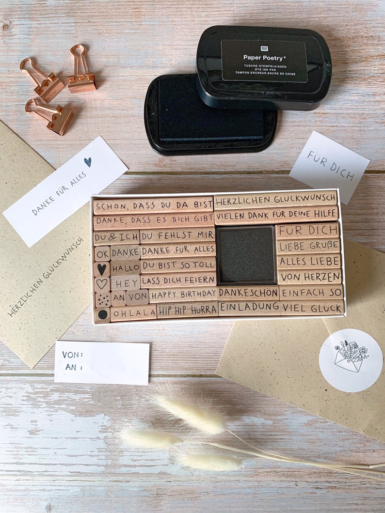 May include: A set of wooden stamps with various phrases in German, including "Danke fur alles", "Herlichen Glueckwunsch", "Du fehlst mir", and "Alles Liebe". The stamps are arranged in a rectangular box with a black ink pad. The box is on a wooden surface with two envelopes and a small white circle with a drawing of an envelope with flowers.
