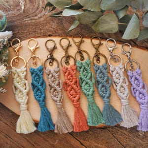 Op de afbeelding: Een verzameling handgemaakte macramé sleutelhangers in verschillende kleuren, waaronder crème, blauw, bruin, koraal, groen en lavendel. Elke sleutelhanger heeft een gevlochten ontwerp en een bijpassende kwast, bevestigd aan een metalen sluiting. De sleutelhangers zijn gerangschikt op een houten oppervlak.