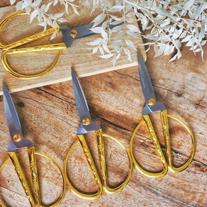May include: Four pairs of gold-colored scissors with ornate designs on a wooden surface. The scissors have silver blades and are arranged in a row.