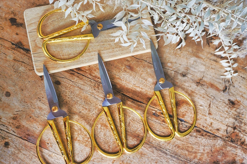 May include: Four pairs of gold-colored scissors with ornate designs on a wooden surface. The scissors have silver blades and are arranged in a row.