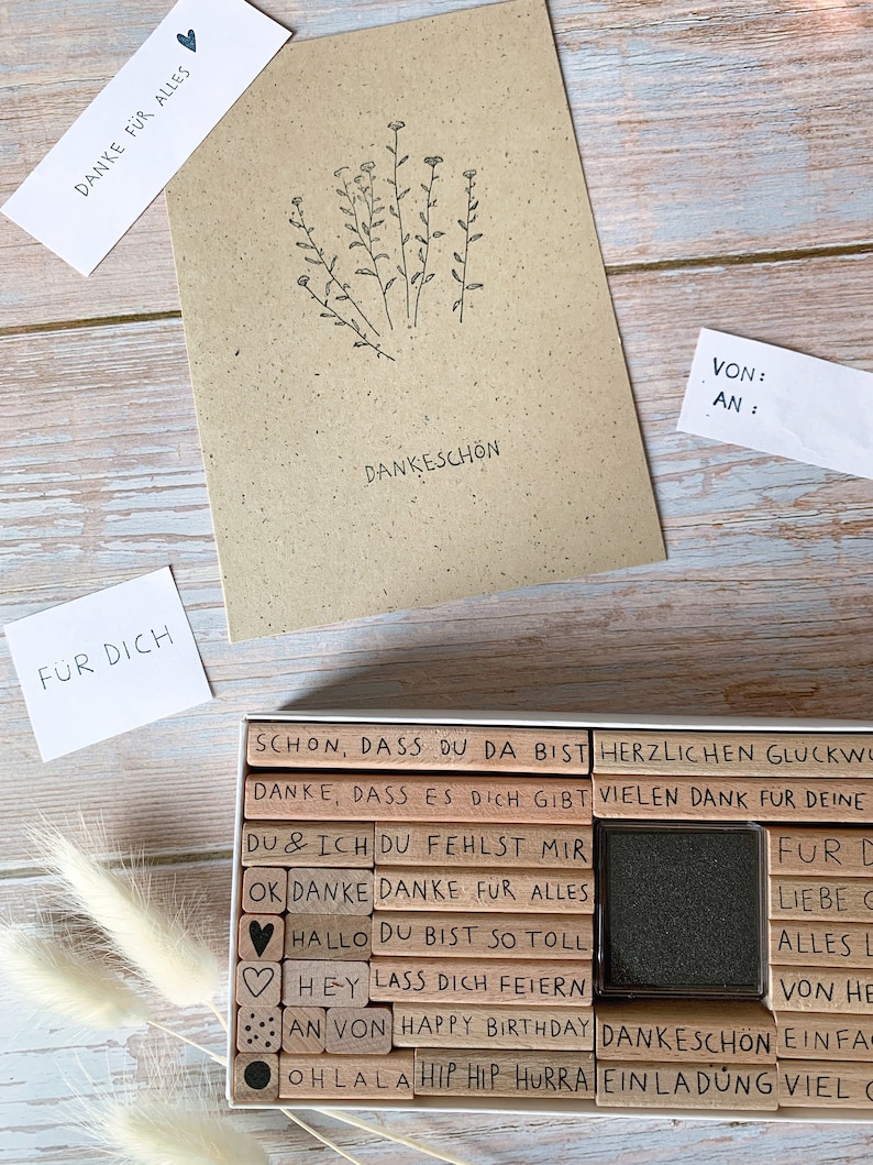 May include: A set of wooden stamps with various phrases and designs, including "Danke", "Happy Birthday", "Oh Lala", and "Herzlischen Gl&uuml;ckwunsch". The stamps are arranged in a wooden box with a black ink pad.