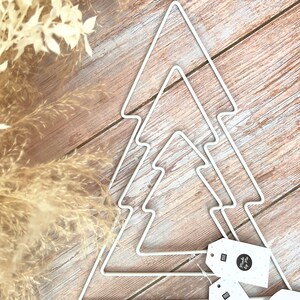 May include: Three white metal Christmas tree shapes on a wood background. The trees are nested inside each other. There are three white tags with black polka dots and the text "Made by Me" on them.