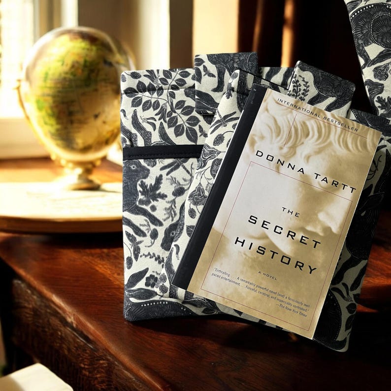 Pode incluir: Um livro intitulado "The Secret History" de Donna Tartt, um best-seller internacional, &eacute; exibido em cima de duas capas de livros com um padr&atilde;o floral preto e branco. As capas dos livros t&ecirc;m uma faixa preta ao redor das bordas.